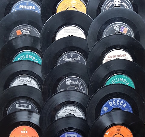 A collection of vintage vinyl records displayed in a pattern. Each record shows a different label from various recording companies such as Columbia, Decca, and Philips. The records have a glossy black finish and the labels feature a mix of colors including blue, orange, and white.