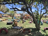 A playground with vibrant, colorful recycled tire structures arranged on the grass, including a swing hanging from a tree. There are tables and chairs set under umbrellas in the background, along with palm trees and clear blue skies, creating a lively outdoor setting.