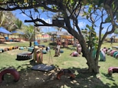 A playground with vibrant, colorful recycled tire structures arranged on the grass, including a swing hanging from a tree. There are tables and chairs set under umbrellas in the background, along with palm trees and clear blue skies, creating a lively outdoor setting.