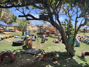 A playground with vibrant, colorful recycled tire structures arranged on the grass, including a swing hanging from a tree. There are tables and chairs set under umbrellas in the background, along with palm trees and clear blue skies, creating a lively outdoor setting.