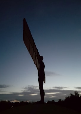 A large angel-like statue silhouetted against a twilight sky with expansive wings stretching upwards. The sky transitions from a deep blue at the top to lighter shades near the horizon, where clouds are lightly scattered. The surrounding area appears dark, with some tree outlines visible at the bottom.