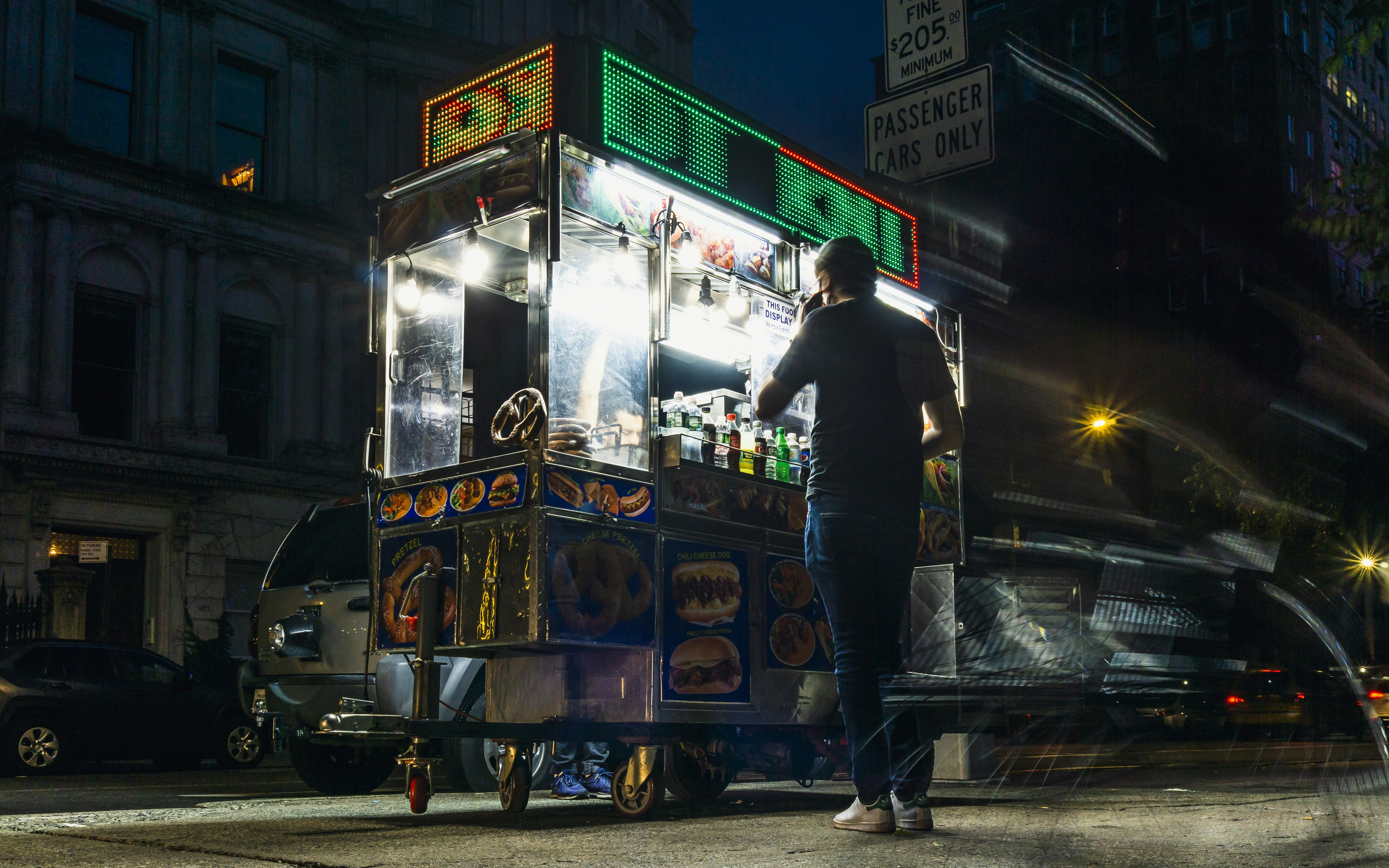 man in black jacket standing beside blue and yellow food cart, Long exposure of a man waiting on his phone getting a hot dog from a street vendor on fifth avenue as a bicycle streaks by.