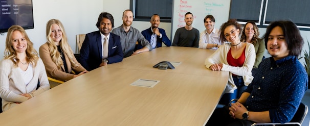 A group of ten people are seated around a long conference table in a well-lit meeting room. They are smiling or posing for the camera, indicating a friendly or professional environment. A whiteboard with scribbles is visible in the background, along with a large screen displaying a list. The table has a central communication device, suggesting a conference or discussion setting.