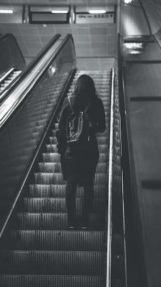 A person with a backpack is ascending an escalator in a dimly lit, monochrome setting. The scene conveys a sense of solitude and movement towards an unknown destination.