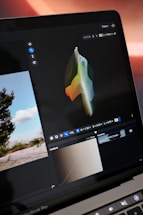 A laptop screen displays a graphics editing program with a colorful 3D shape on a dark background. Various editing tools and settings are visible, including a color curve section, and a small landscape photo in the interface. The scene outside the laptop shows a landscape with trees and a clear sky.