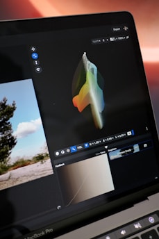 A laptop screen displays a graphics editing program with a colorful 3D shape on a dark background. Various editing tools and settings are visible, including a color curve section, and a small landscape photo in the interface. The scene outside the laptop shows a landscape with trees and a clear sky.