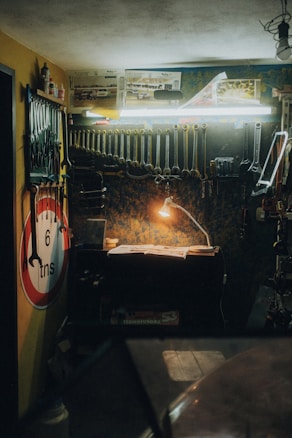 A dimly lit workshop features a wooden workbench illuminated by a small desk lamp. Several tools, including wrenches and pliers, hang meticulously from the wall-mounted rack. A no entry sign and automotive posters adorn the walls, while a toolbox and other workshop essentials are scattered across the bench and shelves.