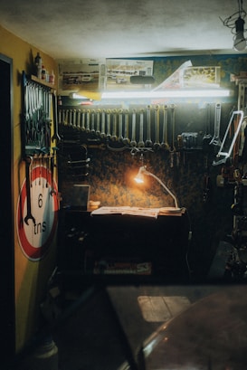 A dimly lit workshop features a wooden workbench illuminated by a small desk lamp. Several tools, including wrenches and pliers, hang meticulously from the wall-mounted rack. A no entry sign and automotive posters adorn the walls, while a toolbox and other workshop essentials are scattered across the bench and shelves.