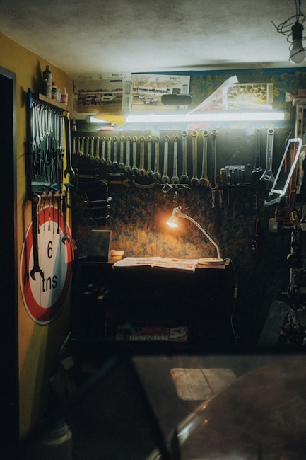 A dimly lit workshop features a wooden workbench illuminated by a small desk lamp. Several tools, including wrenches and pliers, hang meticulously from the wall-mounted rack. A no entry sign and automotive posters adorn the walls, while a toolbox and other workshop essentials are scattered across the bench and shelves.