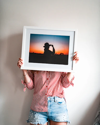 A person wearing a red and white striped shirt and denim shorts holds up a large framed photograph. The photograph within the frame features a silhouette of a person with a hat against a vibrant sunset, blending hues of orange, blue, and black.
