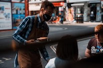 A server wearing a mask, plaid shirt, and apron is delivering food to two seated customers at an outdoor setting, possibly a café or restaurant on a city street. The background shows a blurred view of an urban street with shops and advertising.