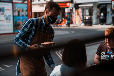 A server wearing a mask, plaid shirt, and apron is delivering food to two seated customers at an outdoor setting, possibly a café or restaurant on a city street. The background shows a blurred view of an urban street with shops and advertising.
