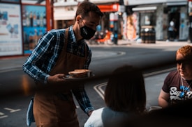A server wearing a mask, plaid shirt, and apron is delivering food to two seated customers at an outdoor setting, possibly a caf&eacute; or restaurant on a city street. The background shows a blurred view of an urban street with shops and advertising.