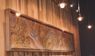 An artistic mural with intricate swirling patterns in a variety of colors is mounted on a wooden wall. It appears to be inspired by traditional or abstract wave designs. Overhead pendant lights are hanging from the ceiling, casting a warm glow.