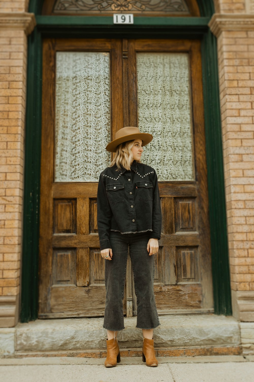 woman in black leather jacket and brown hat standing in front of brown wooden door