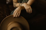 A person with tattooed arms is seated on a dark brown leather couch, holding a brown wide-brimmed hat. Their fingers are adorned with several rings, including a turquoise one, and the tattoos feature floral and geometric designs.