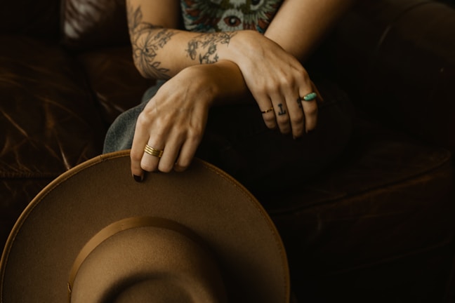 A person with tattooed arms is seated on a dark brown leather couch, holding a brown wide-brimmed hat. Their fingers are adorned with several rings, including a turquoise one, and the tattoos feature floral and geometric designs.