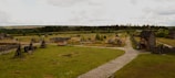 A rustic village setting with wooden structures and fences scattered across a grassy field. The scene includes wooden fortifications, a pathway leading through the area, and several wooden buildings. The landscape is surrounded by forests and fields under a cloudy sky.
