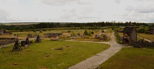 A rustic village setting with wooden structures and fences scattered across a grassy field. The scene includes wooden fortifications, a pathway leading through the area, and several wooden buildings. The landscape is surrounded by forests and fields under a cloudy sky.