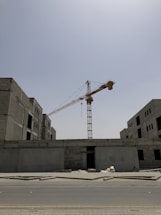 A large construction crane is situated between two unfinished concrete buildings. The crane rises high into a clear sky, and the buildings appear to be in early stages of construction, with exposed brick and unpainted surfaces. A paved road runs in the foreground and there are some construction materials scattered nearby.