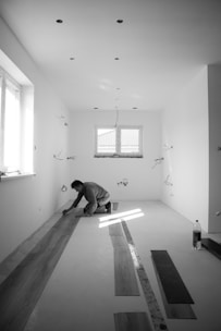 A person is kneeling on the floor in an empty room, placing wooden planks in what seems to be a flooring installation. The room is well-lit by natural light coming through a window. The walls are plain, with a few exposed wires hanging down. There is a bottle placed on the floor, along with several other planks lying around.