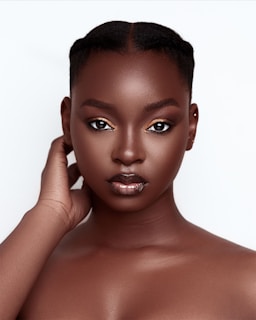 A close-up portrait of a woman with a calm and confident expression. Her skin is smooth and flawless, and her makeup features subtle golden eyeshadow and glossy lips. Her hair is styled neatly, and she touches her neck with one hand.