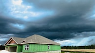 A partially constructed house with green exterior wall panels stands under a dramatic, overcast sky. Dark storm clouds loom, creating a contrast against the lighter patches of blue. The surrounding area features open, undeveloped land with some trees visible on the horizon.