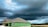 A partially constructed house with green exterior wall panels stands under a dramatic, overcast sky. Dark storm clouds loom, creating a contrast against the lighter patches of blue. The surrounding area features open, undeveloped land with some trees visible on the horizon.