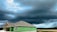 A partially constructed house with green exterior wall panels stands under a dramatic, overcast sky. Dark storm clouds loom, creating a contrast against the lighter patches of blue. The surrounding area features open, undeveloped land with some trees visible on the horizon.