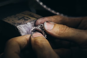 Hands holding and working on a piece of jewelry, possibly a ring, with intricate details and small spikes, against a blurred background of a workbench.