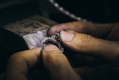 Hands holding and working on a piece of jewelry, possibly a ring, with intricate details and small spikes, against a blurred background of a workbench.