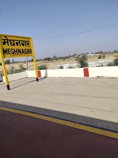 A train station platform with a prominent yellow sign displaying 'MEGHNAGAR' in both Hindi and English. The platform is empty, with a red and yellow painted line along the edge. In the background, there is a clear sky and some fields and trees, giving a rural atmosphere.
