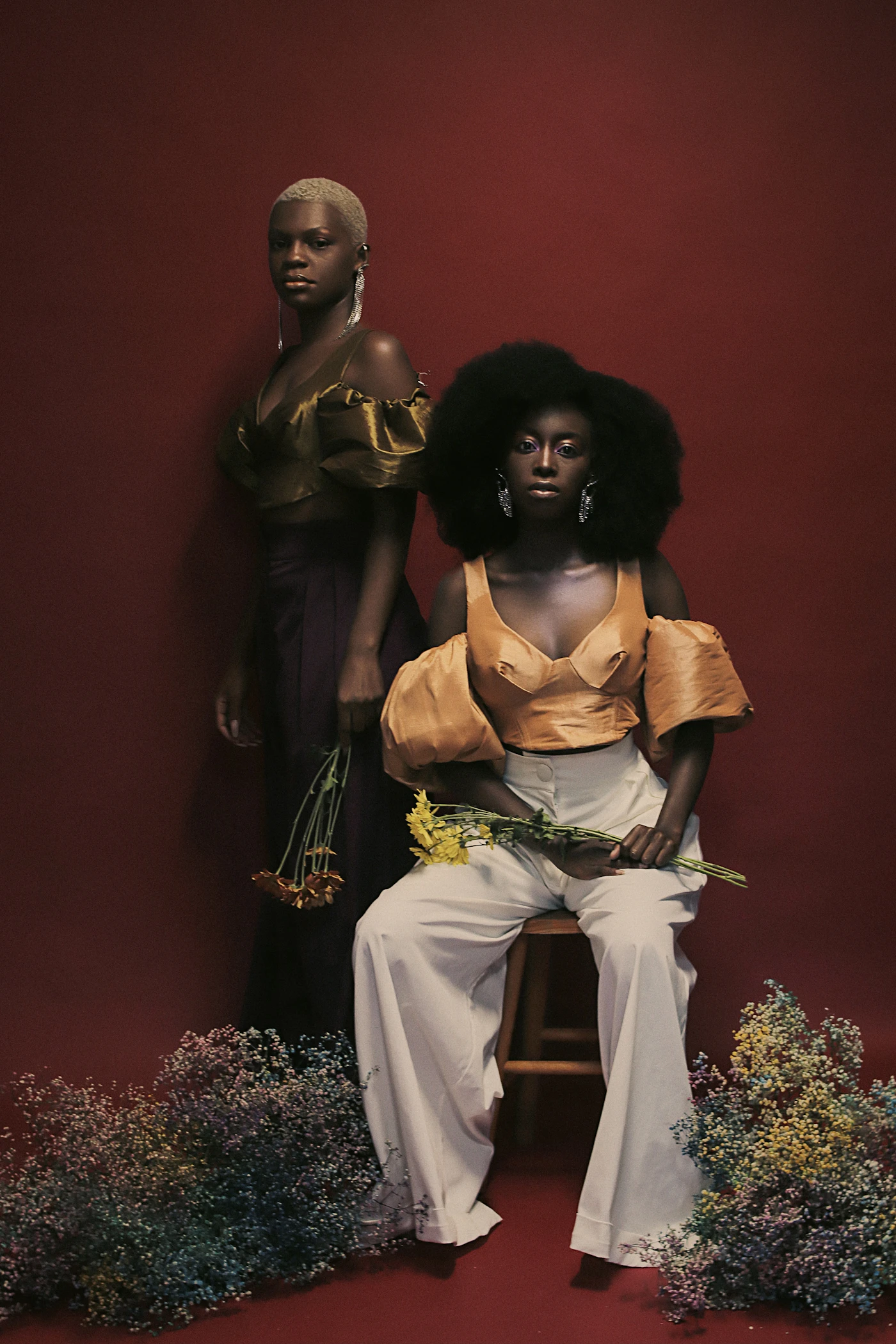 Two models in bronze and apricot silk against a deep red backdrop, surrounded by dried flowers