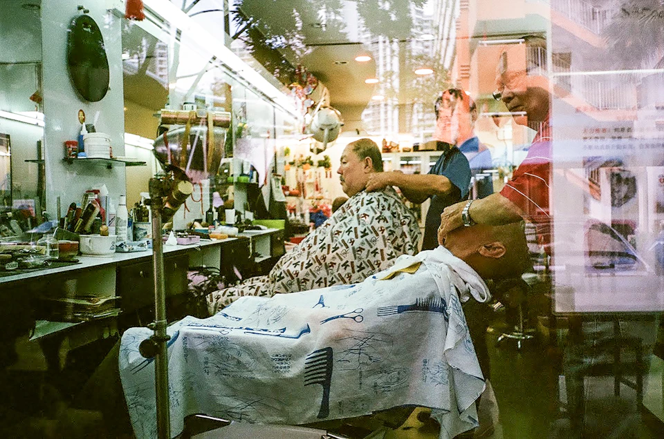 The image depicts a barber shop interior, with three people present - two men and one woman. The man on the left is seated in a chair, wearing a white shirt and a blue and white striped apron. In front of him, another man is sitting in a chair, also wearing an apron. A third person can be seen standing behind them. The barber shop has a window that reflects the interior, adding depth to the scene.