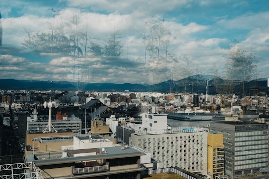 A panoramic view of a cityscape with a mix of modern and traditional architecture, including tall buildings and a pagoda. The city is set against a backdrop of mountains under a partly cloudy sky. The foreground shows reflections of trees on glass, adding an artistic overlay to the scene.