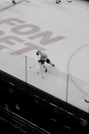 A hockey player in a white uniform is skating on an ice rink, controlling the puck with a hockey stick. Overhead, the ice displays a shadow of large lettering, possibly an advertisement. The player is near the boundary wall on a clean, smooth surface. Other players are partially visible in the distance.