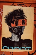 A stylized piece of street art depicts a person with wild hair. The character wears bright orange goggles and a patterned mask, giving a futuristic or rebellious vibe. The background is a textured brown surface with graffiti and the word 'SPORT' partially visible in bold letters.