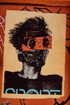 A stylized piece of street art depicts a person with wild hair. The character wears bright orange goggles and a patterned mask, giving a futuristic or rebellious vibe. The background is a textured brown surface with graffiti and the word 'SPORT' partially visible in bold letters.