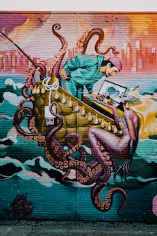 A vibrant mural depicts a surreal scene with a person in a teal outfit interacting with a large octopus. The person appears to be examining or capturing something while being entangled by the octopus's tentacles. In the background, a cityscape can be seen under a dynamically colored sky with hues of orange and pink.