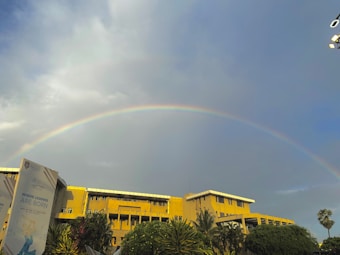 A vibrant rainbow arcs across a partly cloudy sky, with a large yellow building and lush greenery in the foreground. A poster with the text 'WHERE LEGENDS ARE BORN' is visible on the left. The lighting suggests a serene, possibly late afternoon setting.