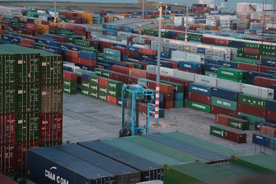 A vast array of colorful shipping containers stacked in rows at a busy port. A large blue crane is visible, facilitating the movement of the containers. The scene suggests an industrial setting with a backdrop of water and more containers in the distance.