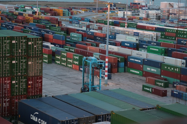A vast array of colorful shipping containers stacked in rows at a busy port. A large blue crane is visible, facilitating the movement of the containers. The scene suggests an industrial setting with a backdrop of water and more containers in the distance.