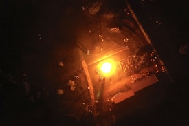 An aerial view capturing a central, bright bonfire illuminating its surroundings. The fire is situated in an open area, with people forming a circle around it, casting visible shadows. Adjacent structures and pathways are faintly visible in the low-lit environment, creating a sense of gathering and warmth.