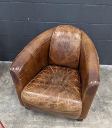 A vintage leather armchair with a worn and distressed finish sits on a concrete floor against a dark brick wall. The chair is rounded with a high back and armrests, showing visible creases and texture.