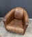 A vintage leather armchair with a worn and distressed finish sits on a concrete floor against a dark brick wall. The chair is rounded with a high back and armrests, showing visible creases and texture.