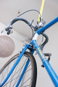 A close-up view of a vintage blue bicycle with a yellow and white logo. The bike features drop handlebars wrapped in grey tape and shiny gear shifters on the downtube. Part of a woven light fixture is in the background.