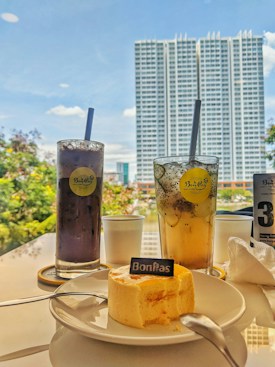 A table is set with two tall glasses of drinks, one dark purple and the other light yellow with visible fruit bits. A small cake with a 'BonPas' label is placed on a white plate with a spoon. The background features a large building with numerous windows against a bright blue sky and hints of green foliage.