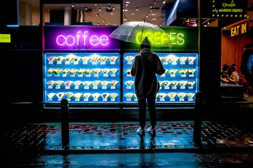 A person stands under an umbrella in front of a brightly lit display of plastic food items, specifically crepes, outside a cafe. The neon signs in pink and green read 'coffee' and 'crepes', respectively. Reflections of the lights shimmer on the wet pavement, suggesting recent rain. Inside the cafe, a few people are visible, sitting and dining.