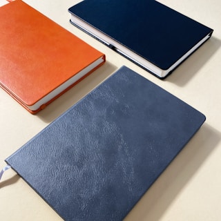 Three hardcover notebooks with textured covers are arranged on a light surface. The notebooks are in different colors: orange, blue, and grey.