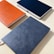 Three hardcover notebooks with textured covers are arranged on a light surface. The notebooks are in different colors: orange, blue, and grey.
